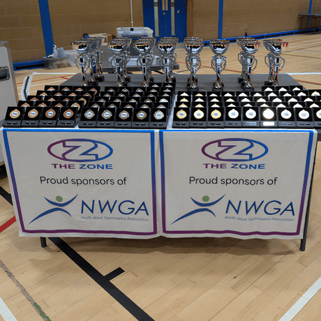 Prize medals for gymnastics competition on a table, with a banner reading 'The Zone, Proud Sponsors of NWGA'